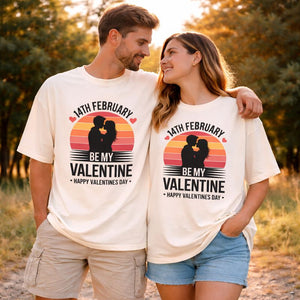 Verliebtes Paar in warmem Sonnenlicht trägt cremefarbene Oversized T-Shirts mit Retro-Sonnenuntergang und Silhouetten-Motiv „Be My Valentine – Happy Valentine’s Day“, romantisches Valentinstags-Design von Bright Light Arts.