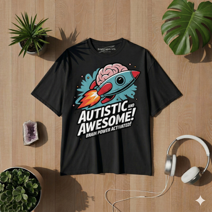 Draufsicht auf ein schwarzes Oversized-T-Shirt, das flach auf einer Holzoberfläche liegt. Auf der Brust befindet sich ein farbenfroher Grafikdruck im Comic-Stil, der eine hellblaue Rakete mit einem rosa Gehirn an der Spitze zeigt. Darunter steht in fetten weißen Großbuchstaben der Text "AUTISTIC AND AWESOME!" und kleiner darunter "BRAIN POWER ACTIVATED!". Das T-Shirt ist von verschiedenen Requisiten umgeben: Oben links eine Topfpflanze, unten links Sukkulenten und ein violetter Kristallstein, rechts ein wei