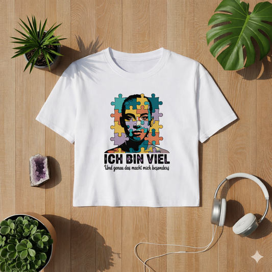 Weißes Cropped Damen T-Shirt mit „Ich bin viel“ Autismus Puzzle Portrait Design als Flatlay auf hellem Holzboden mit Pflanzen und Kopfhörern – modernes Lifestyle Mockup