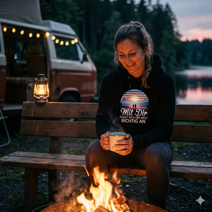 Frau im kuscheligen schwarzen Bright Lights Arts Hoodie mit atmosphärischem Bergsee-Print am Lagerfeuer.