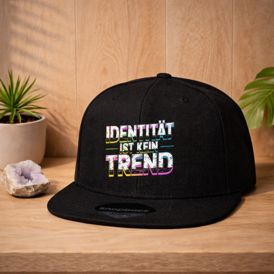 Schwarze Snapback Cap mit „Identität ist kein Trend“ Design im Bright Lights Arts Stil, präsentiert als Lifestyle Mockup auf hellem Holz mit Pflanzen und ruhiger Atmosphäre