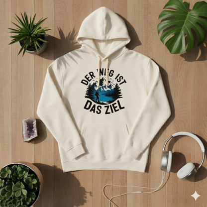 Draufsicht auf einen creme-farbenen Kapuzenpullover auf hellem Holzboden, bedruckt mit einem runden Motiv, das Berge, einen See, einen Wanderer und den deutschen Text „DER WEG IST DAS ZIEL“ zeigt. Der Hoodie ist umgeben von Sukkulenten, einem Monstera-Blatt, einem Amethysten und weißen Kopfhörern