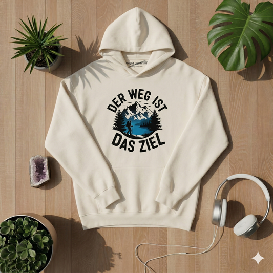Draufsicht auf einen creme-farbenen Kapuzenpullover auf hellem Holzboden, bedruckt mit einem runden Motiv, das Berge, einen See, einen Wanderer und den deutschen Text „DER WEG IST DAS ZIEL“ zeigt. Der Hoodie ist umgeben von Sukkulenten, einem Monstera-Blatt, einem Amethysten und weißen Kopfhörern