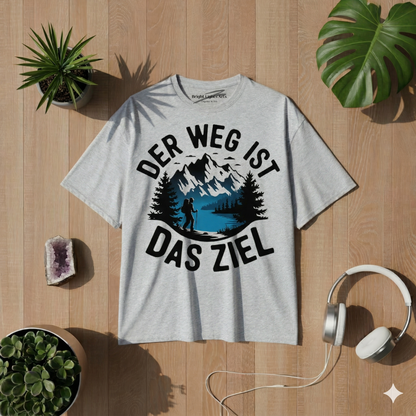 Draufsicht auf ein graumeliertes Oversized-T-Shirt mit einem runden Print, der Berge, einen See und einen Wanderer zeigt, umgeben von dem Text "DER WEG IST DAS ZIEL". Das T-Shirt liegt auf einem hellen Holzboden, flankiert von Monstera-Blättern, Sukkulenten, einem Amethysten und weißen Kopfhörern.