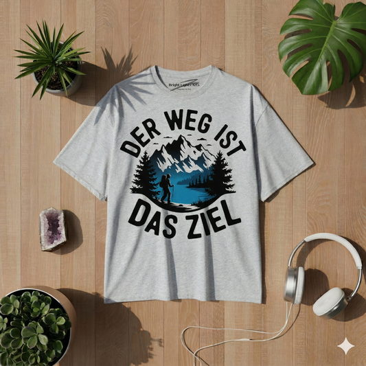 Draufsicht auf ein graumeliertes Oversized-T-Shirt mit einem runden Print, der Berge, einen See und einen Wanderer zeigt, umgeben von dem Text "DER WEG IST DAS ZIEL". Das T-Shirt liegt auf einem hellen Holzboden, flankiert von Monstera-Blättern, Sukkulenten, einem Amethysten und weißen Kopfhörern.
