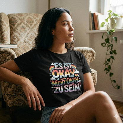 Authentische junge Frau in einem gemütlichen Zimmer, die das 'Es ist okay' Shirt trägt und entspannt nachdenkt.