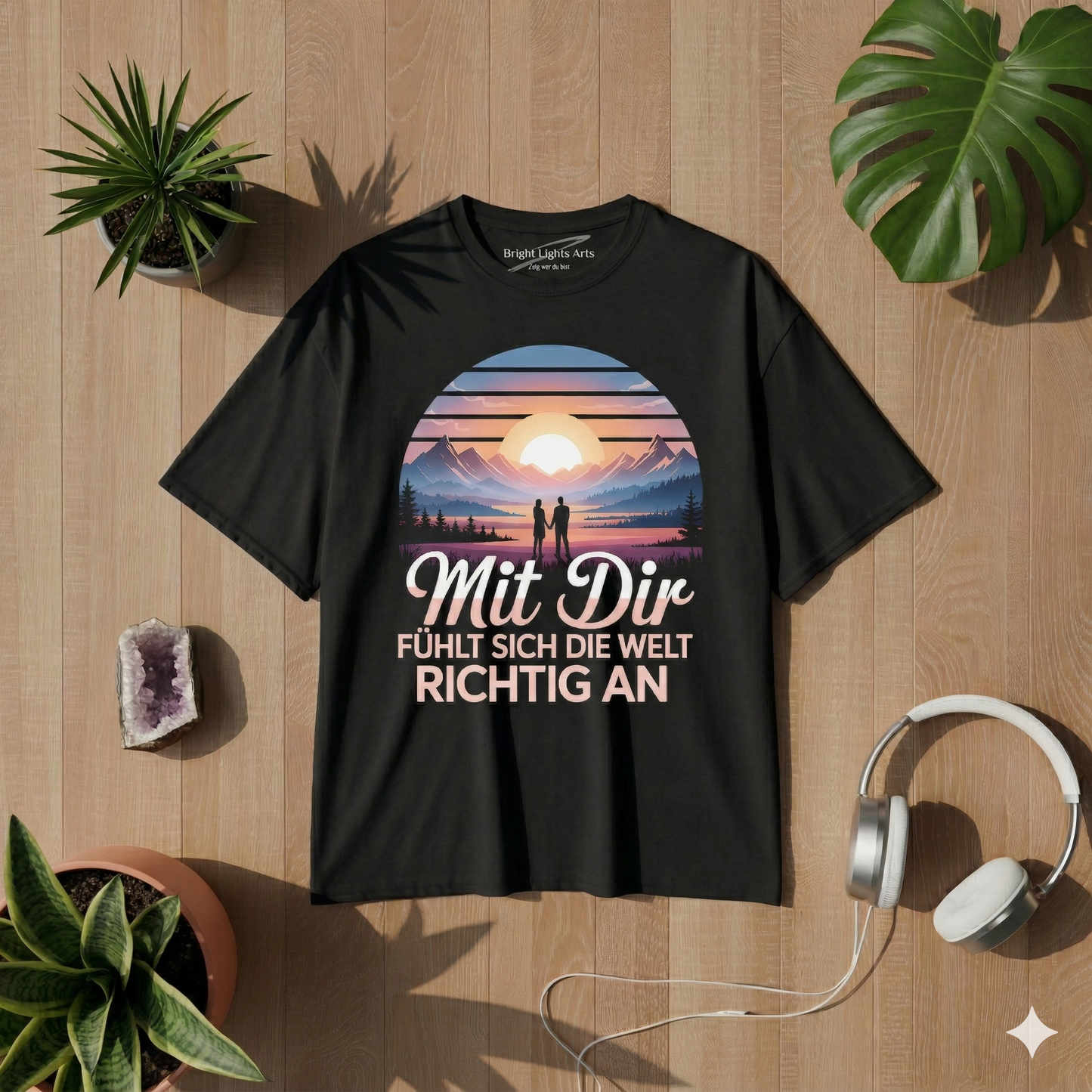 Draufsicht auf ein schwarzes Oversized-T-Shirt aus Bio-Baumwolle mit dem Grafik-Print „Mit Dir“, der ein verliebtes Paar vor einem Sonnenuntergang in den Bergen zeigt. Das T-Shirt liegt auf einem hellen Holzboden, umgeben von einem Monstera-Blatt, einem Sukkulententopf, einer Amethystdruse und weißen Kopfhörern. Der Drucktext lautet: „Mit Dir FÜHLT SICH DIE WELT RICHTIG AN“. Das Nackenetikett zeigt das Branding „Bright Lights Arts“.