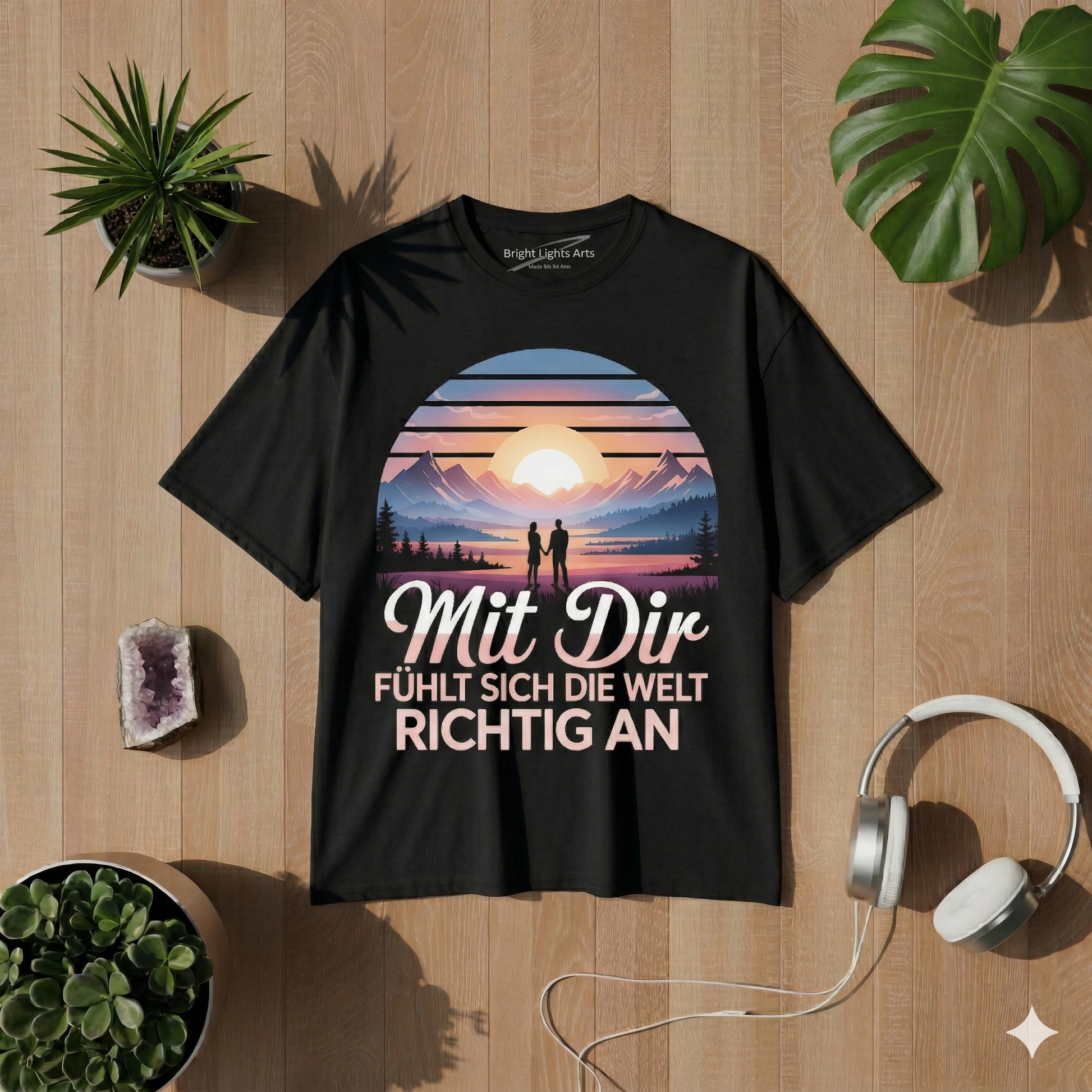 Draufsicht auf ein schwarzes oversized T-Shirt mit dem "Mit Dir fühlt sich die Welt richtig an" Design, umgeben von einem Amethyst-Kristall, weißen Kopfhörern, zwei Sukkulenten-Pflanzen und Monstera-Blättern auf einem hellen Holzhintergrund bei sonniger Lichtstimmung