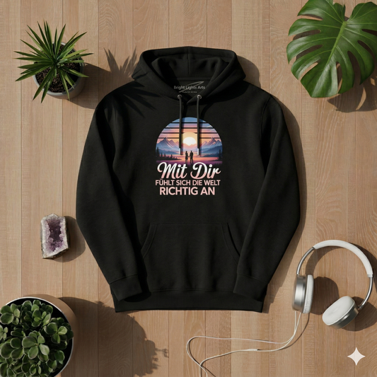 Draufsicht auf ein schwarzes oversized T-Shirt mit dem "Mit Dir fühlt sich die Welt richtig an" Design, umgeben von einem Amethyst-Kristall, weißen Kopfhörern, zwei Sukkulenten-Pflanzen und Monstera-Blättern auf einem hellen Holzhintergrund bei sonniger Lichtstimmung.